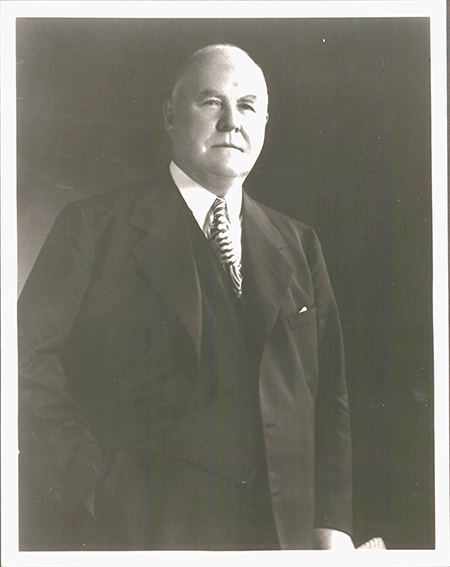 Elderly man in a suit and tie, standing with a serious expression in a black and white photograph.