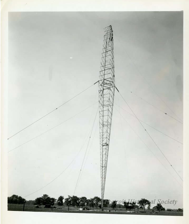 Black and white photo of the construction of the WJR radio antenna. Black and white photo of the construction of the WJR antenna outside of the WJR Radio Transmitting Building on the southwest corner of Sibley Road and Grange Road in Riverview. Several construction workers and piles of building material are scattered around the base of the tower.