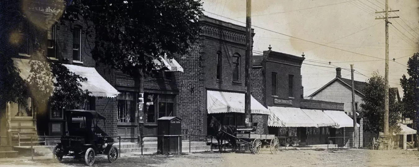 Historic street scene with vintage buildings, cars, and horse-drawn wagon under an overcast sky.