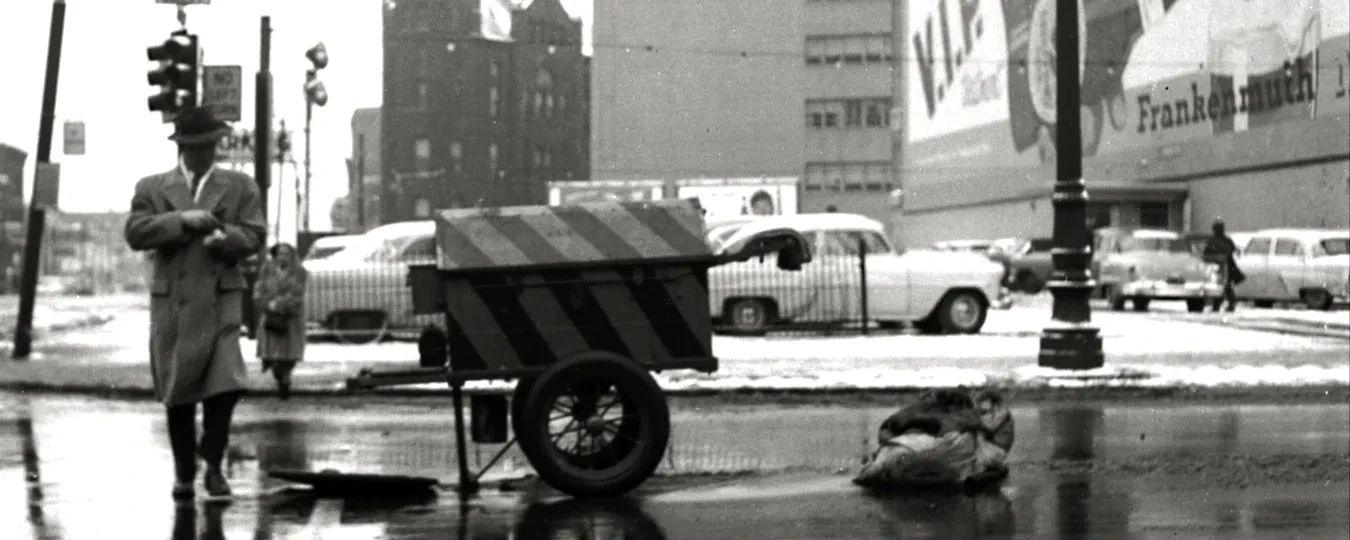 Man next to a cart on a wet, snowy city street corner, tall buildings and billboard in background, black and white photograph.