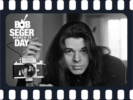Black and white photo of a man with long hair in a dark turtleneck next to a camera on a tripod. with a filmstrip border and the words Bob Seger Day March 15