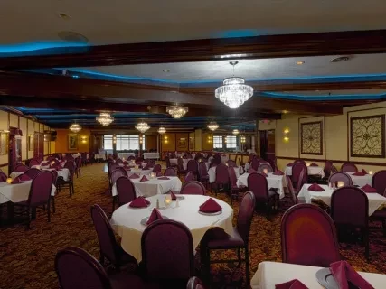 Elegant dining room with round tables set with white cloths and red napkins, under chandeliers and warm lighting.