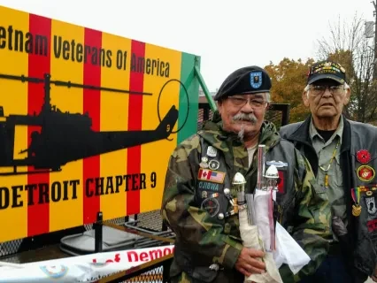 Two elderly veterans in decorated jackets stand beside a Vietnam Veterans of America sign outdoors.