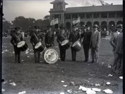 Band of uniformed men with drums and flags on a grassy field, with a large building and scattered papers in the background.