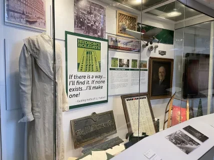 Museum display with historical artifacts, including a dress, framed posters, photographs, and a portrait.