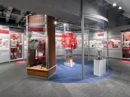 Hockey museum exhibit with red jerseys displayed in glass cases, trophies, and historical photos on walls.