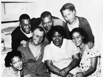 Group of seven people, three seated, four standing behind, smiling and gathered closely, in black and white.