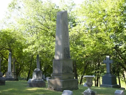 Gravestones in a cemetery under a canopy of green trees on a sunny day.