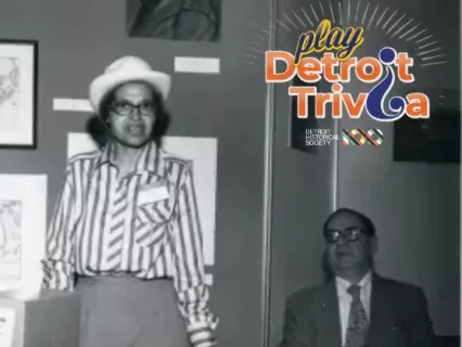 Three men in vintage attire, one standing between two seated, maps on the wall; retro setting.