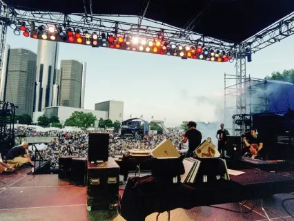 Outdoor concert stage with musicians, city skyline in the background, and a crowd gathered below.