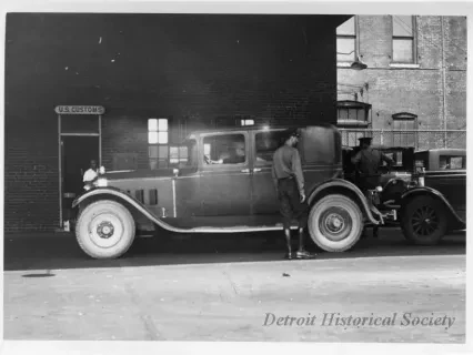 Black and white photo of a vintage car parked in front of a brick building with two people standing beside it.