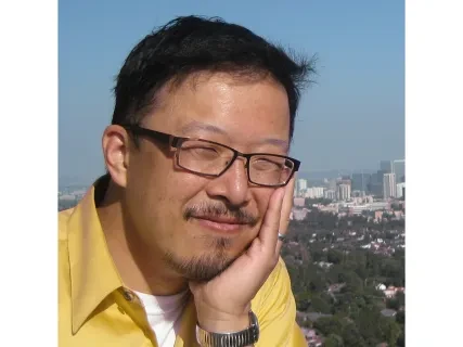 Man in glasses smiling outdoors with city skyline in the background.