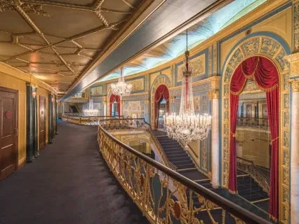 Ornate theater interior with chandeliers, red curtains, and gold detailing along a curved balcony.