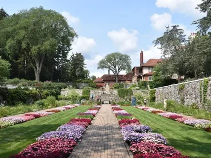 Formal garden with colorful flower beds, stone pathway, large trees, and a brick house in the background under a blue sky.