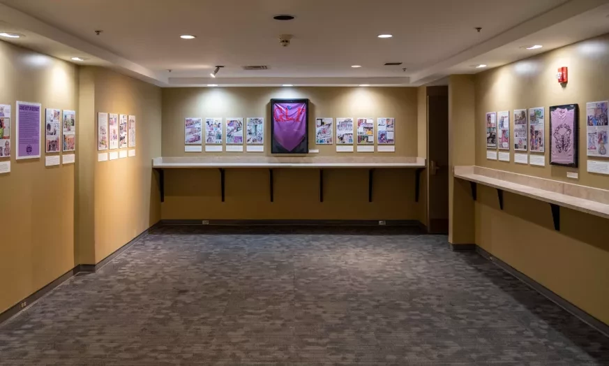 Art gallery with framed pictures on beige walls, focused lighting, and carpeted floor. Central artwork features a large purple shape.