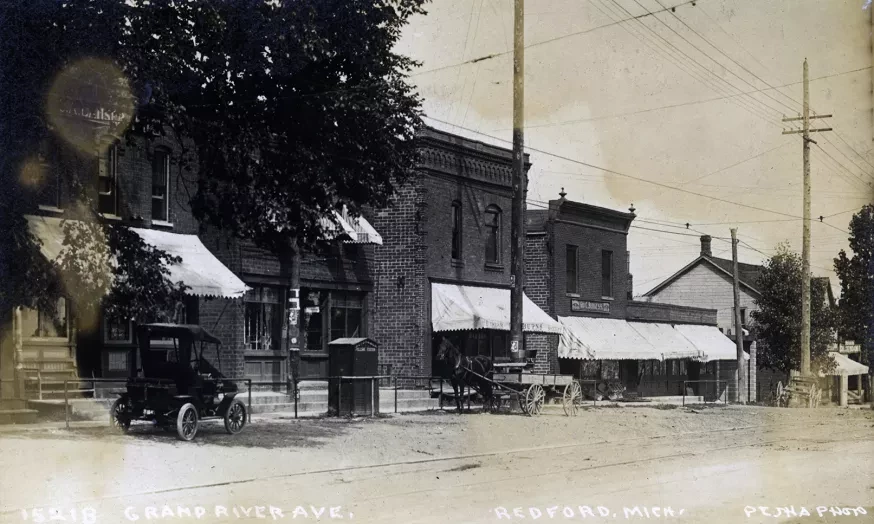 Historic street scene with vintage buildings, cars, and horse-drawn wagon under an overcast sky.