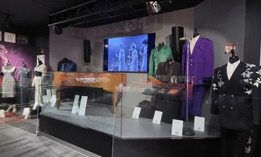 Museum display of vintage mannequins in colorful suits behind glass, with a screen showing historical footage.