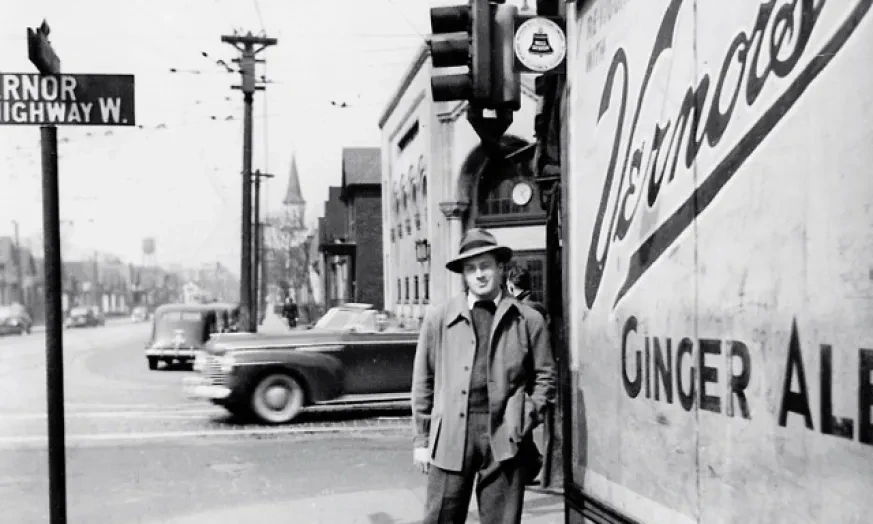 Man in coat and hat standing by a Vernors Ginger Ale sign at a street corner with passing car in the background.