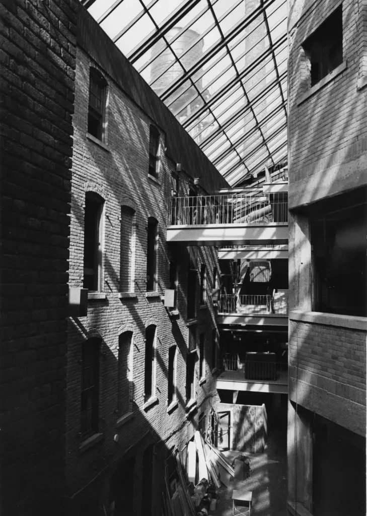 Glass-roofed atrium with brick walls and a narrow walkway, sunlight casting geometric shadows.