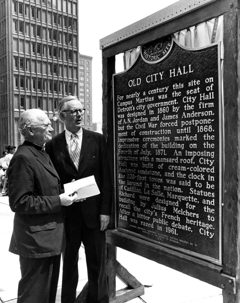 Two men in suits stand beside a historical marker for Old City Hall, discussing the text. A tall building is in the background.