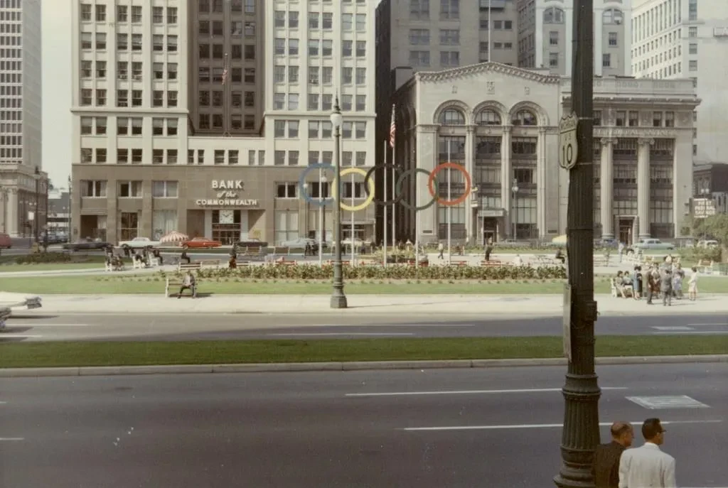 Street view of a park with people gathered, surrounded by tall buildings and a lamp post in the foreground.