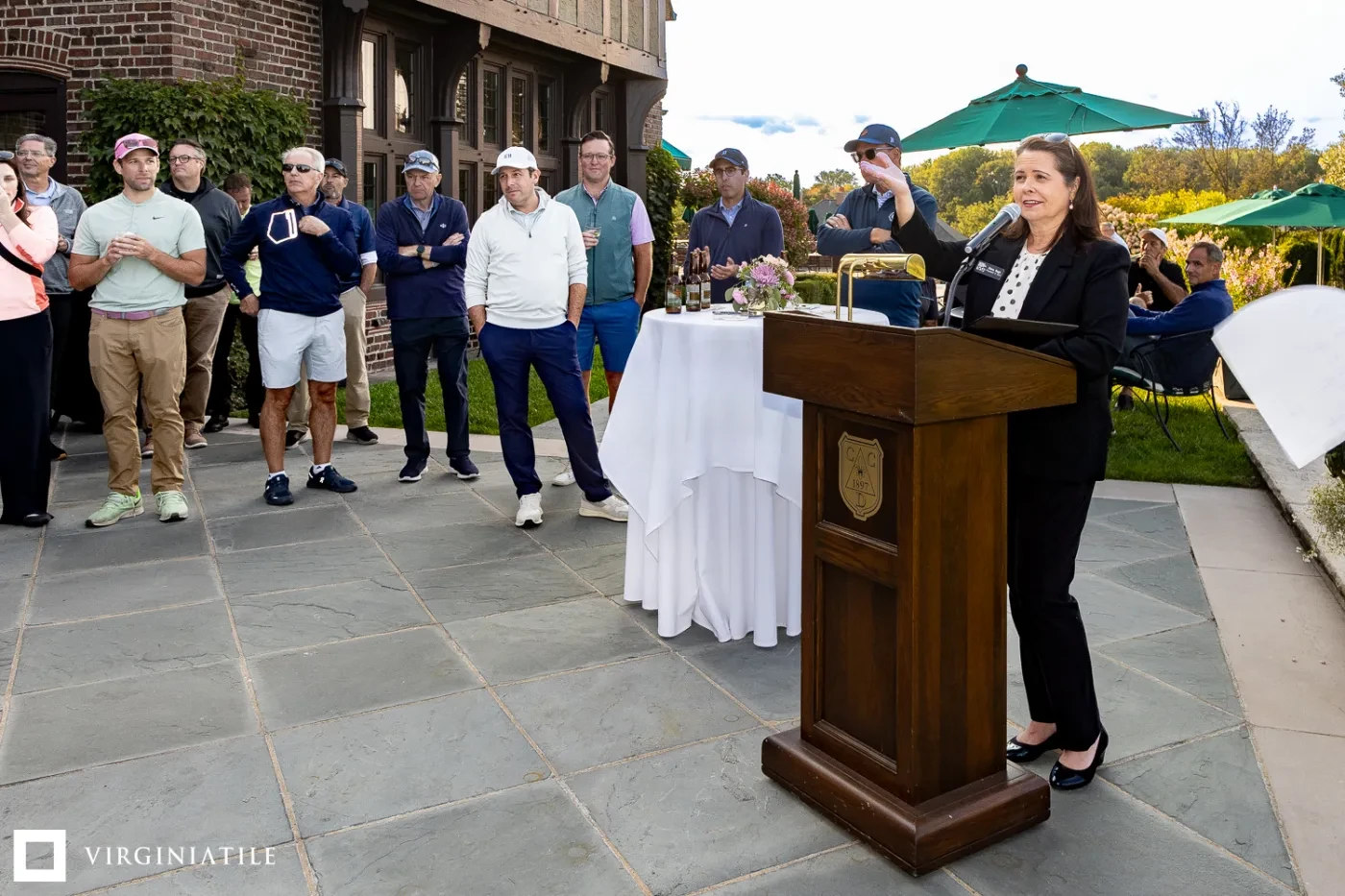 Woman speaking at a podium outdoors with a group of people listening nearby under green umbrellas.