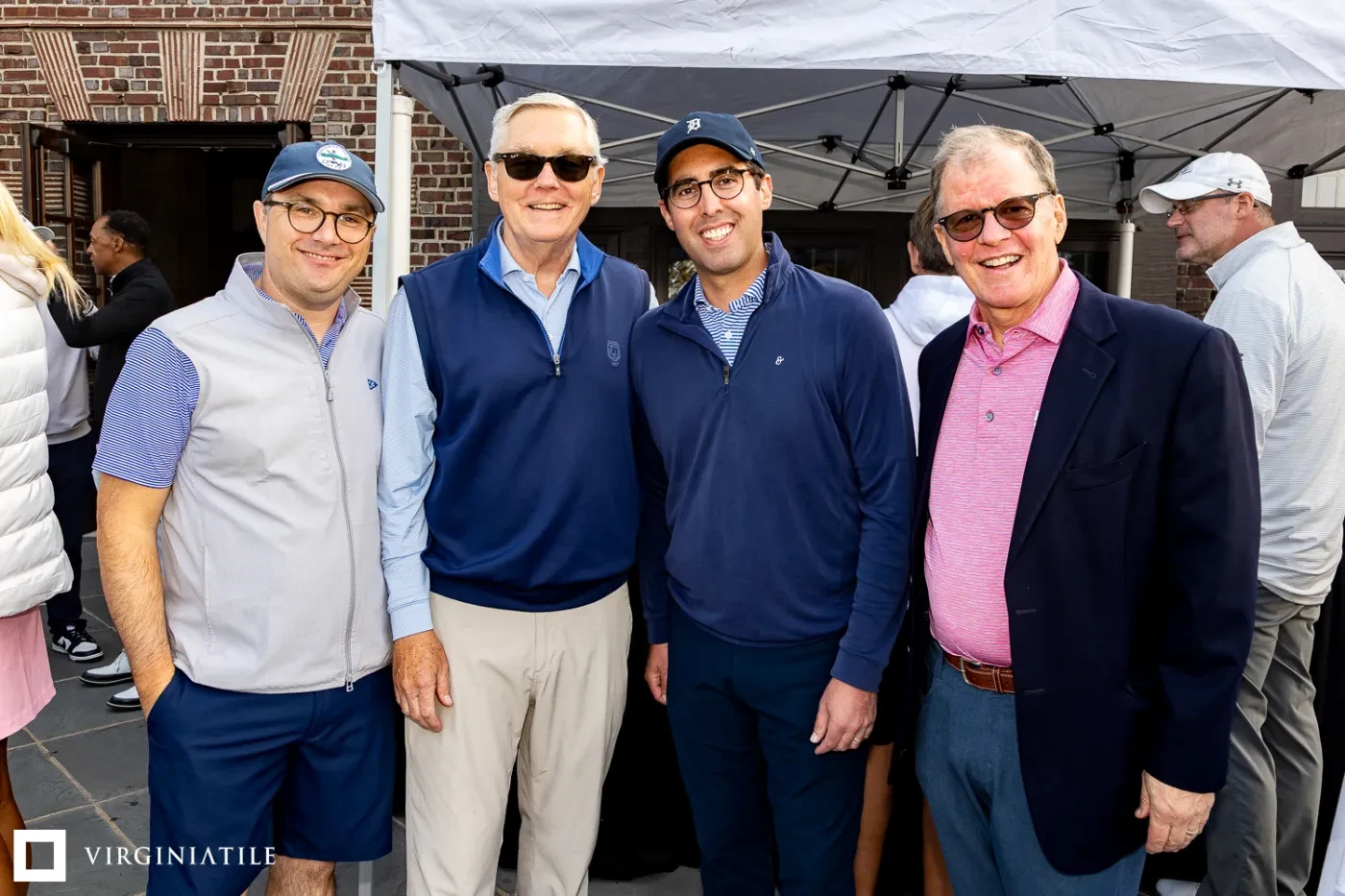 Four men smiling outdoors under a white tent, dressed in casual and semi-formal attire, brick wall in background.