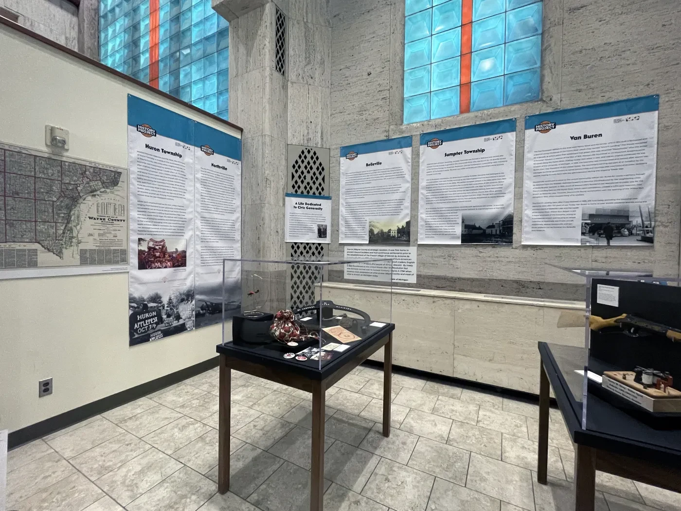 Museum exhibit room with informational panels on walls, display tables with artifacts, and blue-tinted windows.