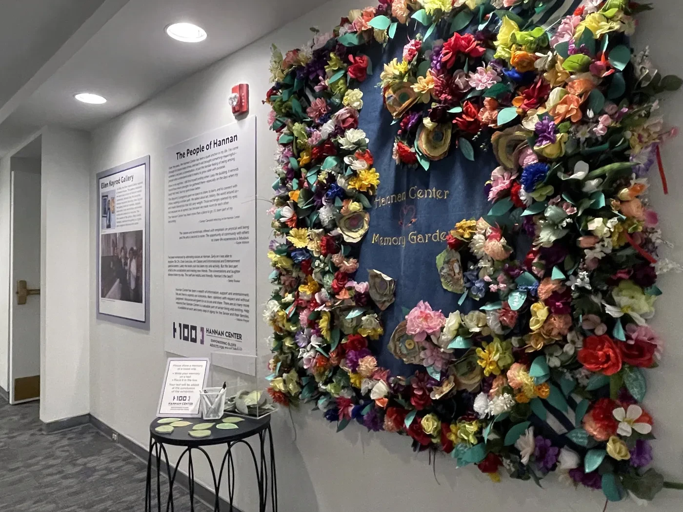 A colorful floral tapestry on a white hallway wall, with informational posters and a small table nearby.