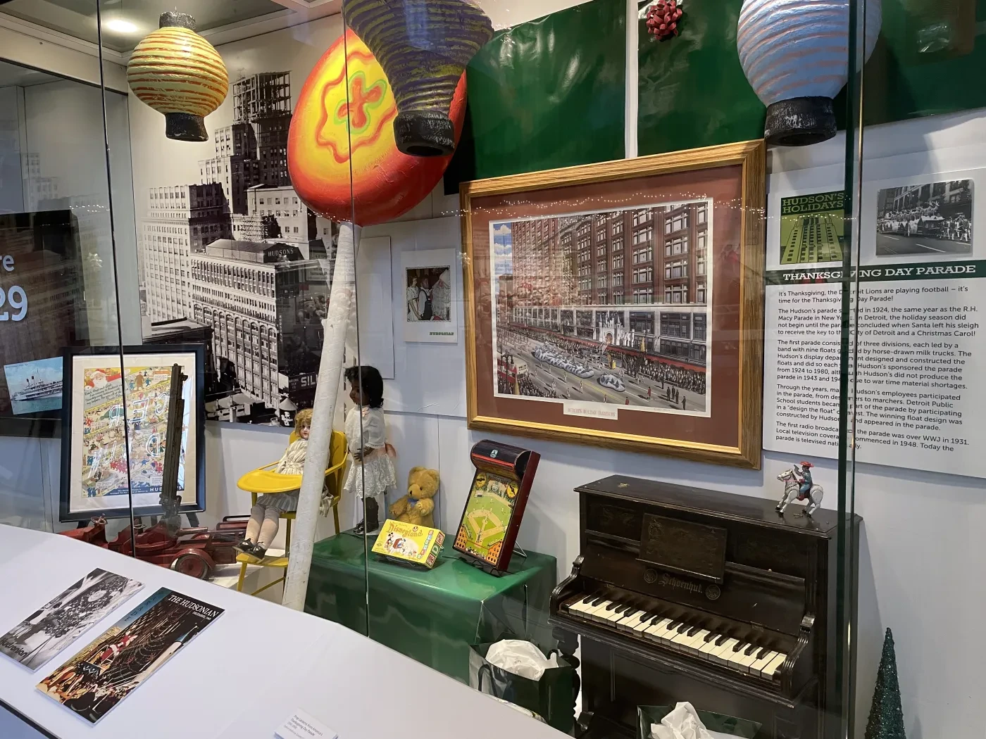 Museum display with a framed city photo, colorful lanterns, a small piano, and cultural artifacts in a glass case.
