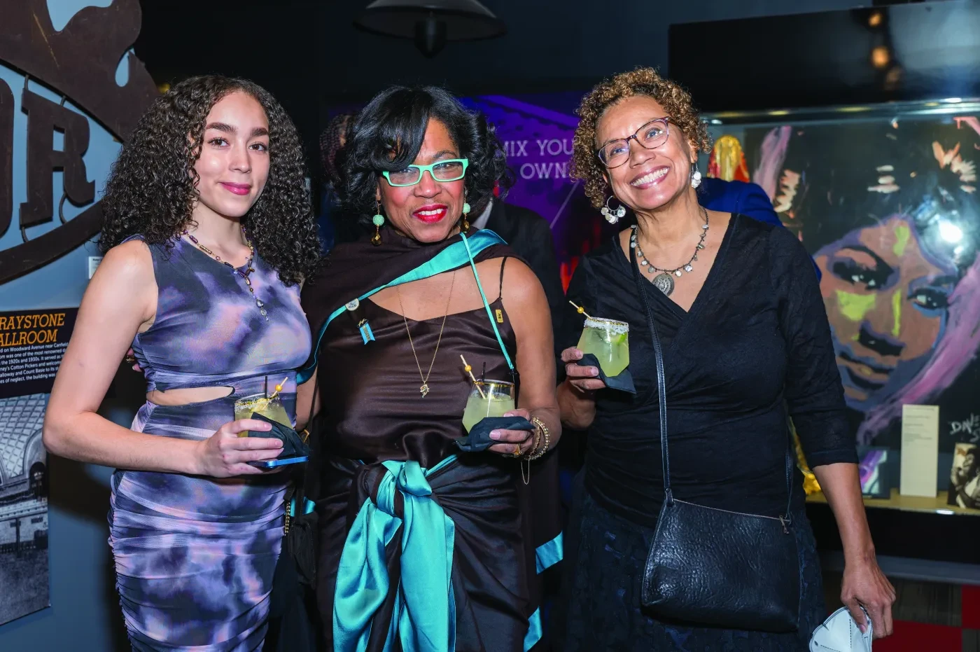 Three women smiling and holding drinks at an indoor event, with colorful wall art in the background.