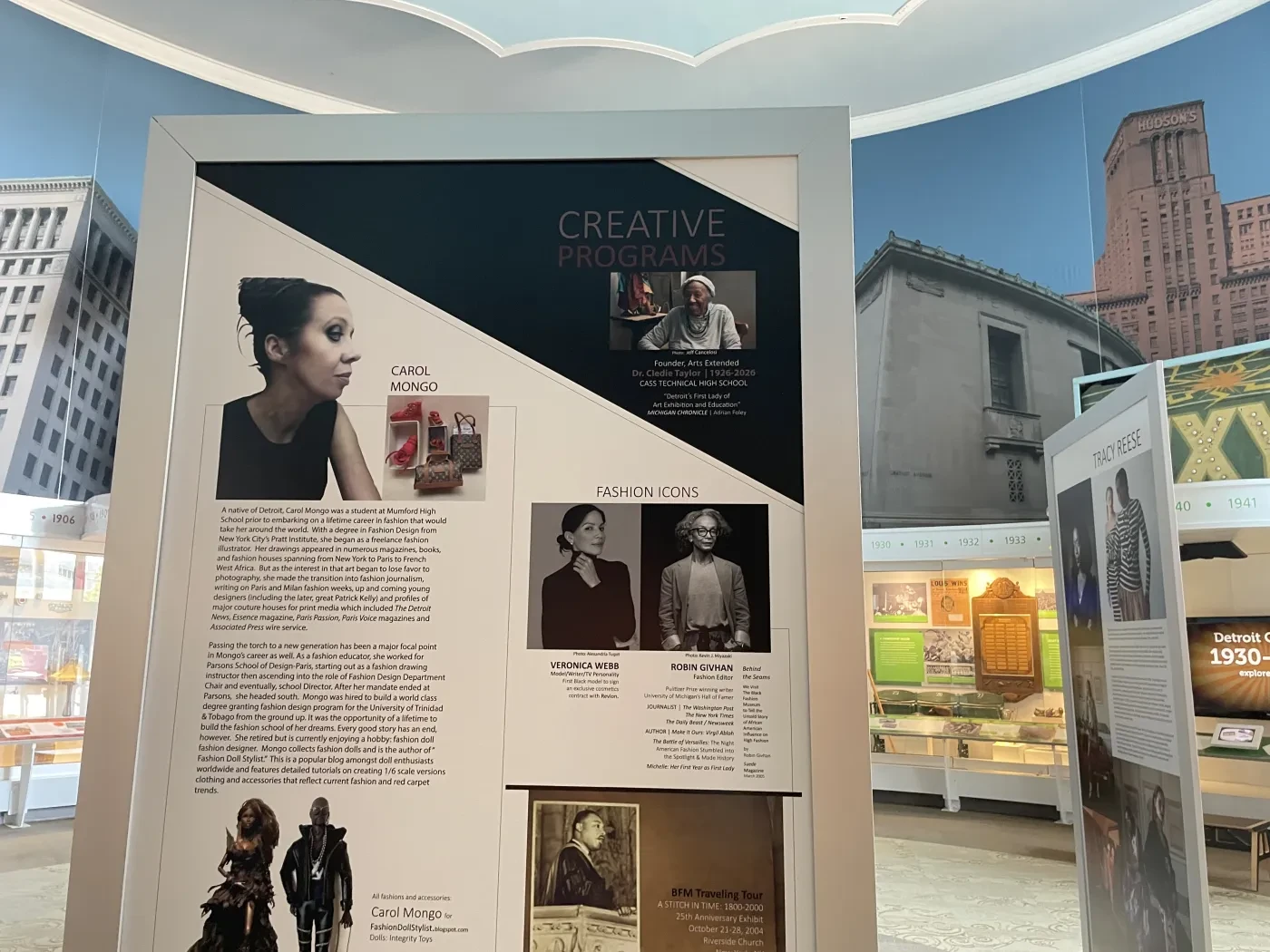 Museum exhibit display with photographs, text, and images in a room with a cityscape background.