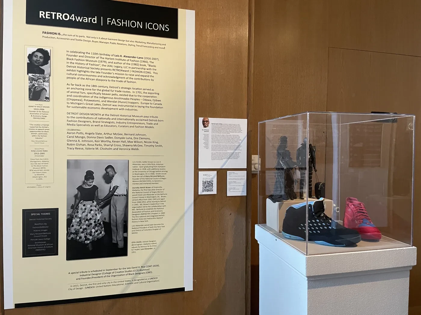 Exhibit display with fashion-themed text panel and mannequins in colorful garments inside a glass case.