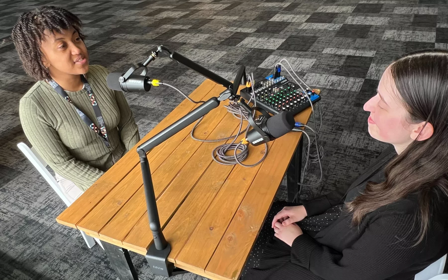 Two women recording a podcast at a wooden table in a studio with microphones and audio equipment.