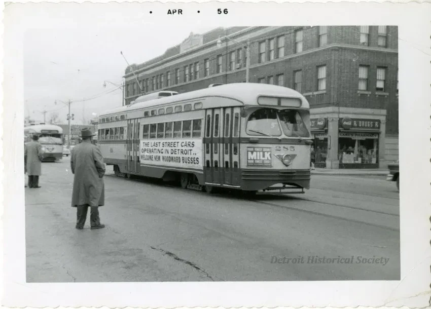 Streetcar on city road, person in overcoat crossing, vintage buildings and another streetcar in background, black and white photo.