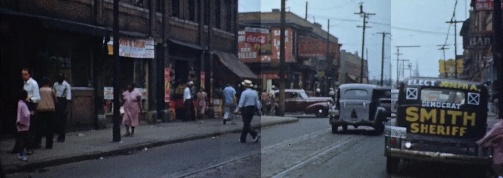 Street scene with vintage cars, pedestrians, and a "Smith Sheriff" sign on a vehicle. Buildings line the road.