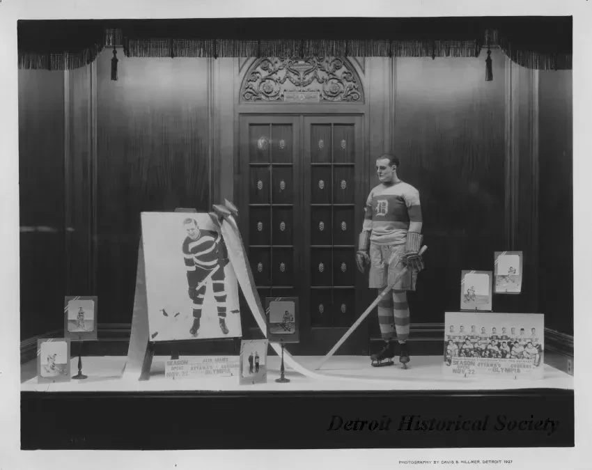 Hockey player mannequin in vintage uniform next to a framed portrait and photographs in a wood-paneled room.