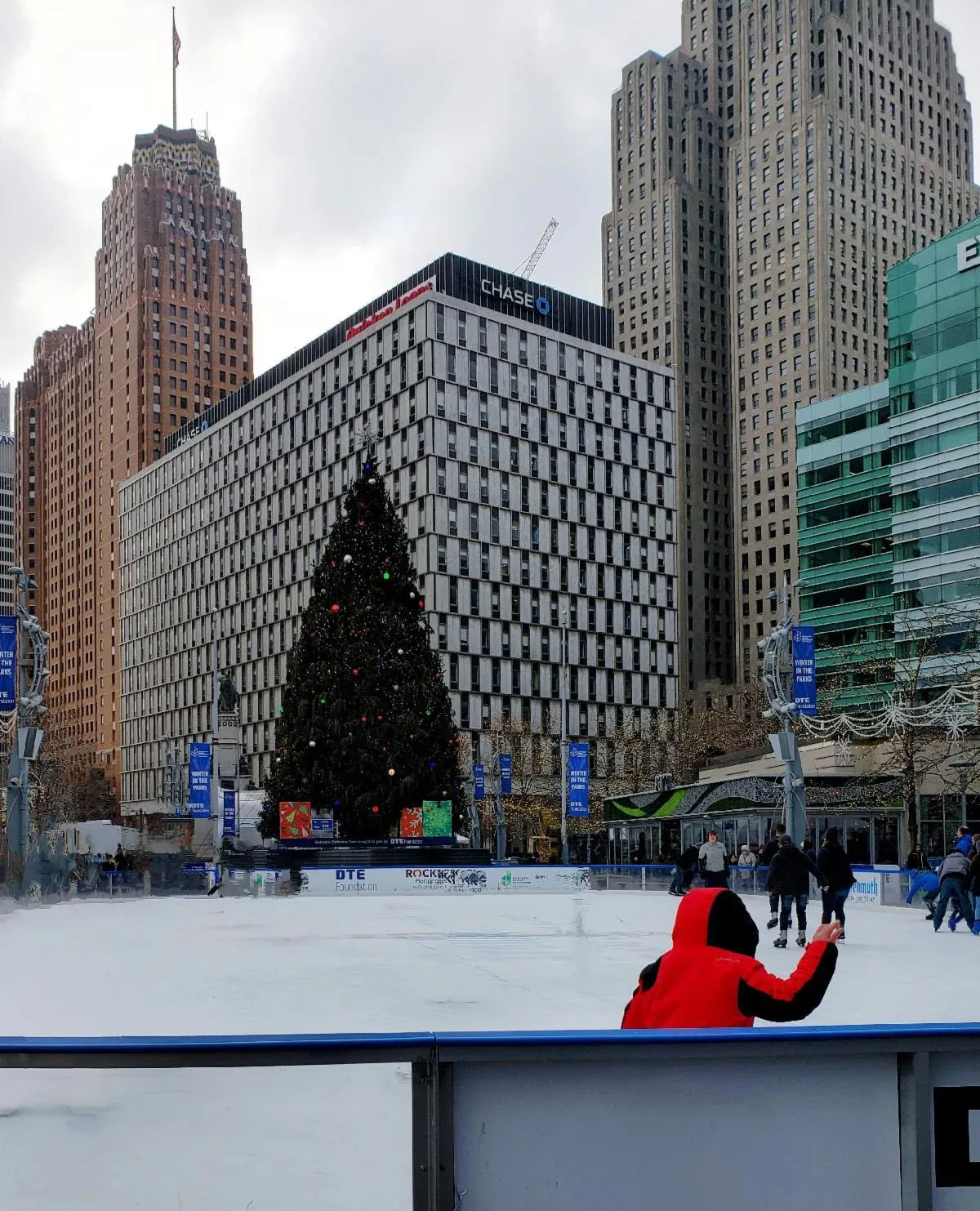 Skaters on an ice rink with a large Christmas tree, surrounded by tall buildings on a cloudy day.