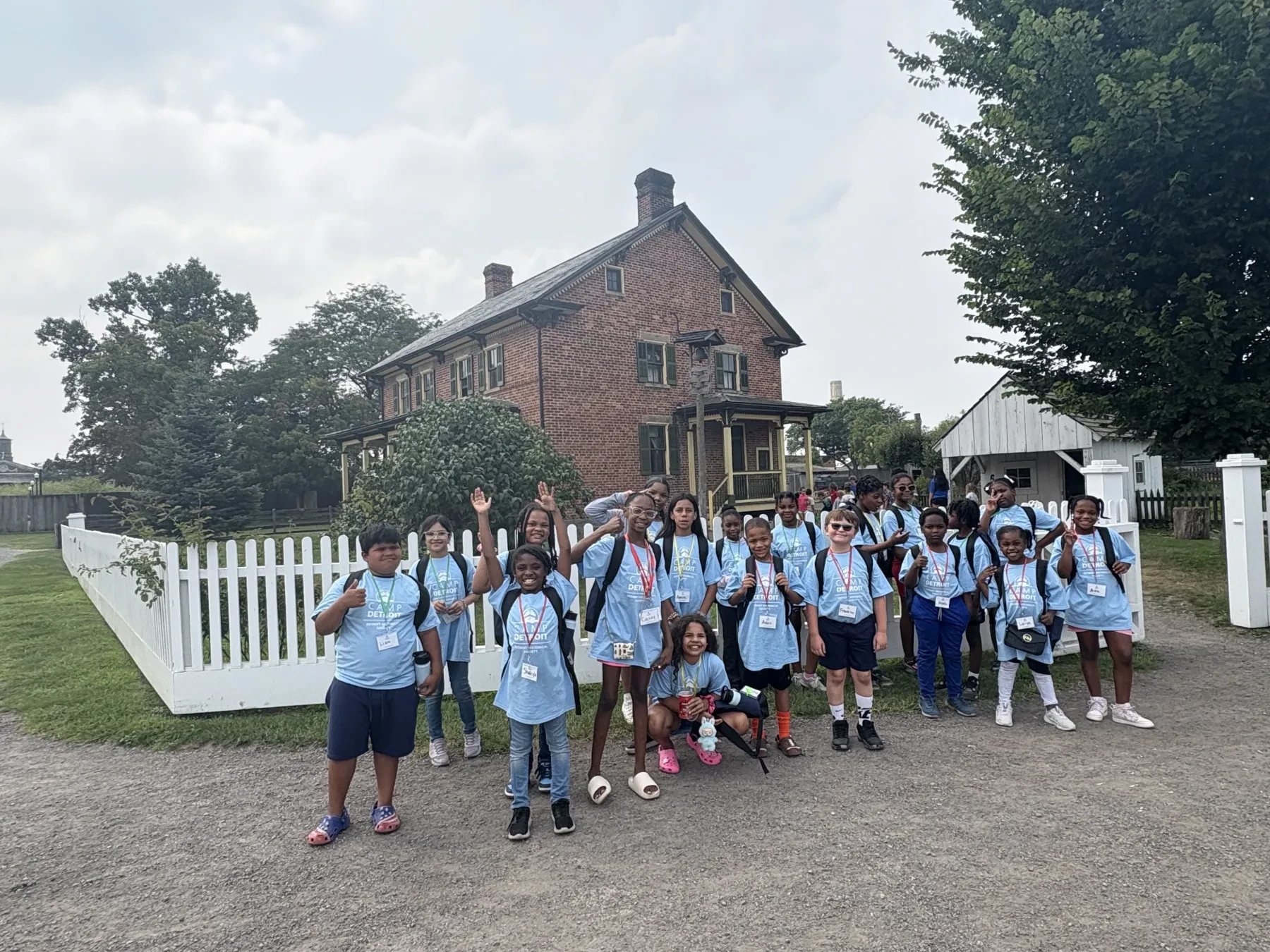 A group of children in blue shirts stands outside a brick house with white fencing on a cloudy day.