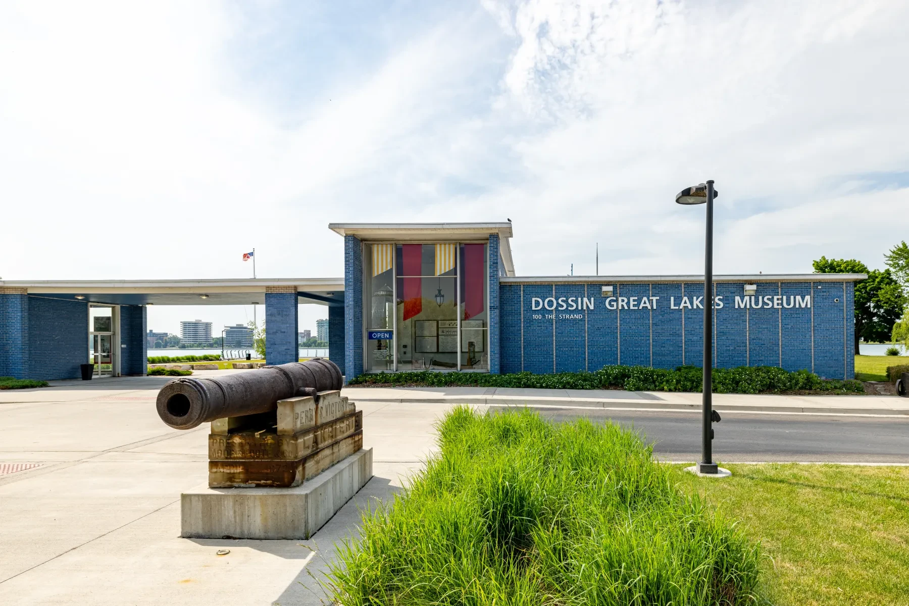 Cannon displayed outside a modern museum with a blue facade and large windows, surrounded by greenery.